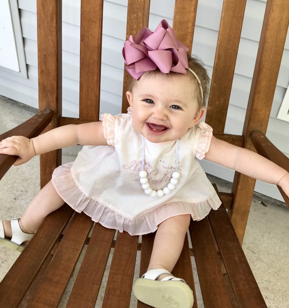 A baby girl sitting on a wooden chair with a pink bow.