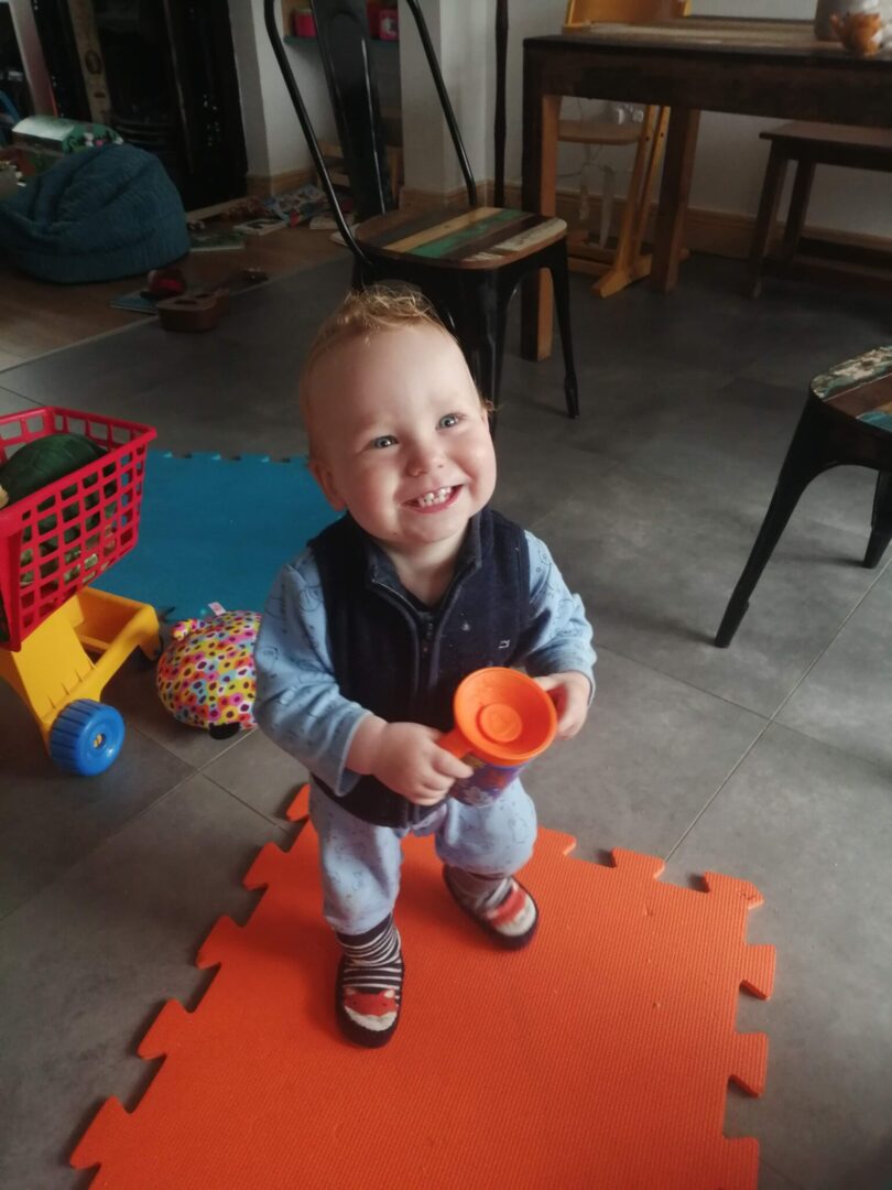 A baby standing on an orange mat with a cup.