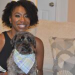 A woman is sitting on a couch with a dog in a bandana.