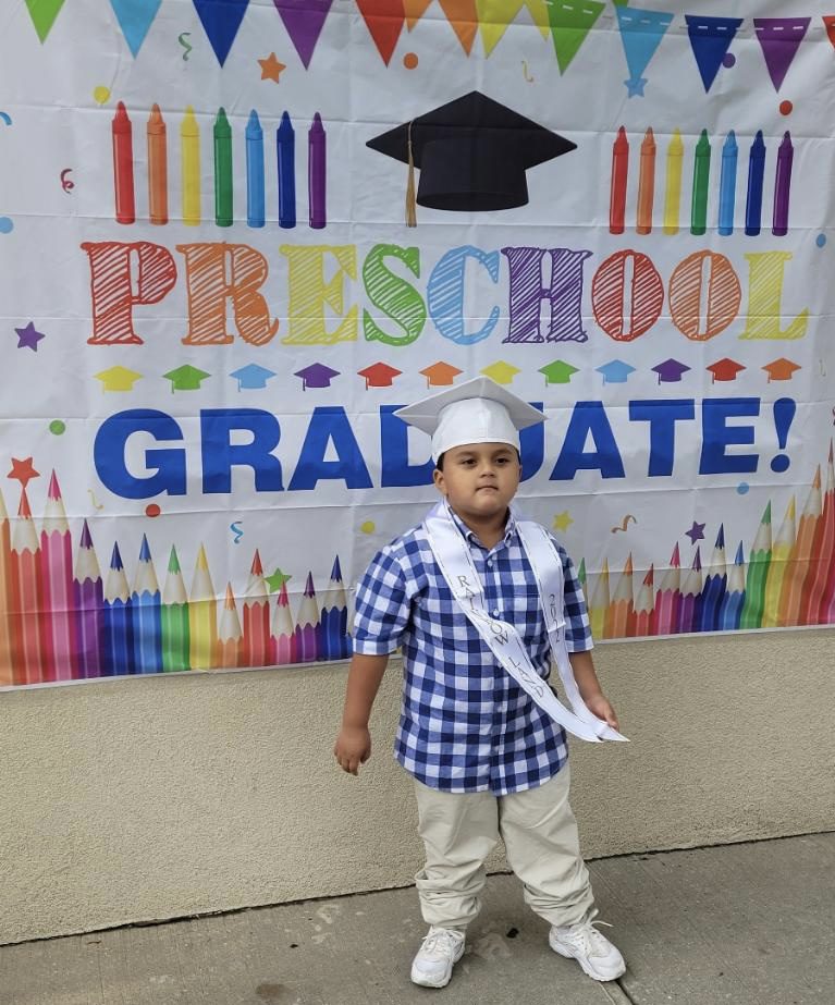 A boy standing in front of a banner that says preschool graduate.