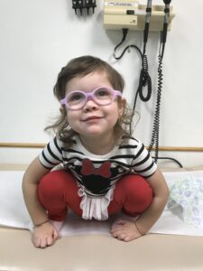 A little girl wearing glasses and sitting on a bed.