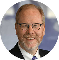 A man wearing glasses and a suit smiles.