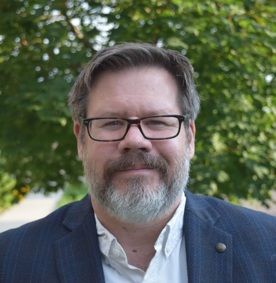 A man with glasses and a beard standing in front of a tree.