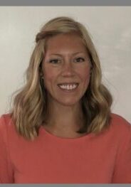 A woman in an orange shirt is smiling in front of a white wall.