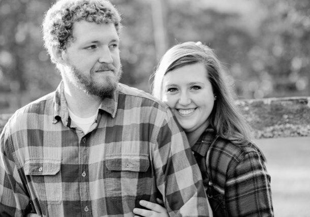 A black and white photo of a couple posing in front of a fence.