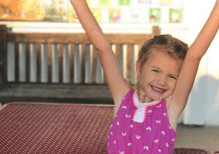 A little girl is sitting on a table with her arms raised.