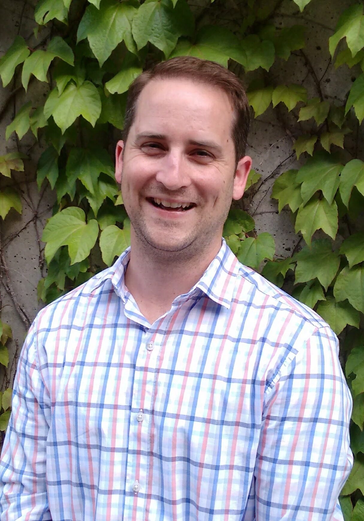 A man in a plaid shirt standing in front of an ivy covered wall.