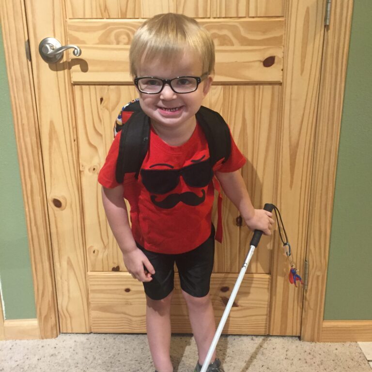 A young boy with glasses and a cane standing in front of a door.