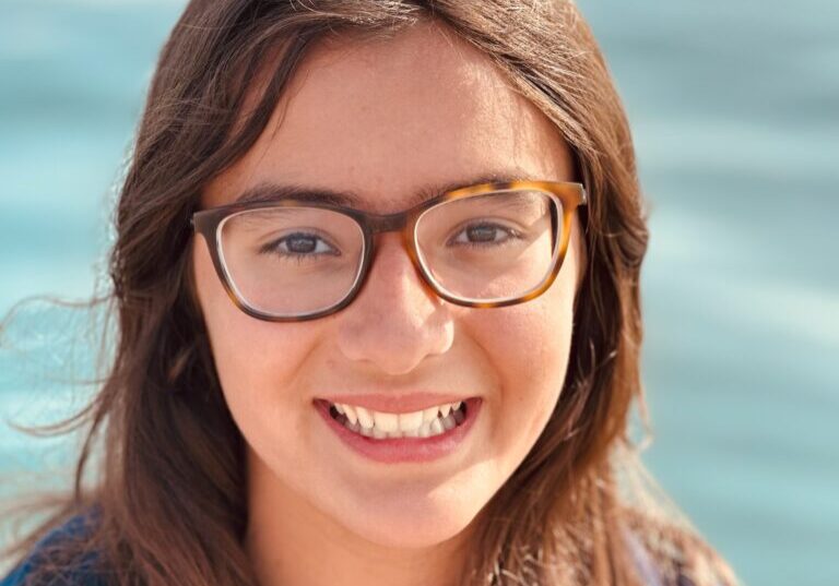 A young girl wearing glasses smiles in front of the water.