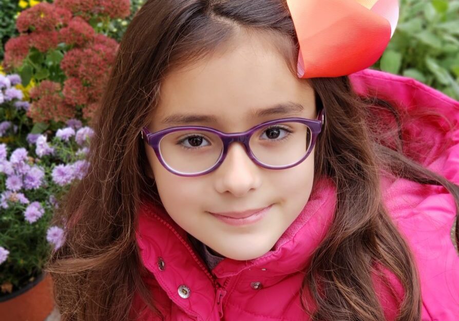 A little girl wearing glasses and a rainbow bow.