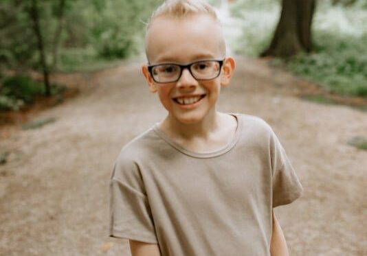 A young boy wearing glasses and a t - shirt in a wooded area.