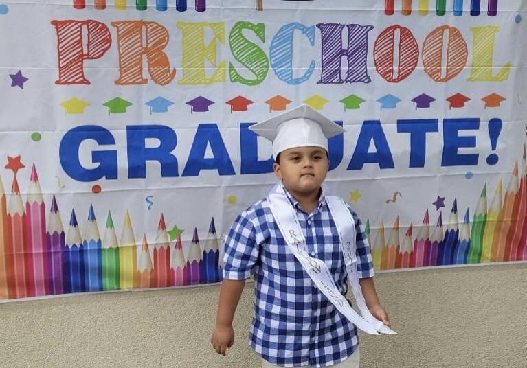 A boy standing in front of a banner that says preschool graduate.