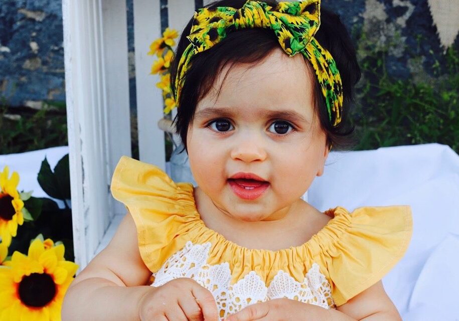 A baby girl wearing a yellow dress and sunflower headband.