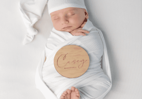 A newborn baby wrapped in a white blanket with a wooden name plaque.