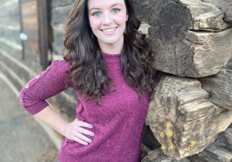 A young woman leaning against a log cabin.