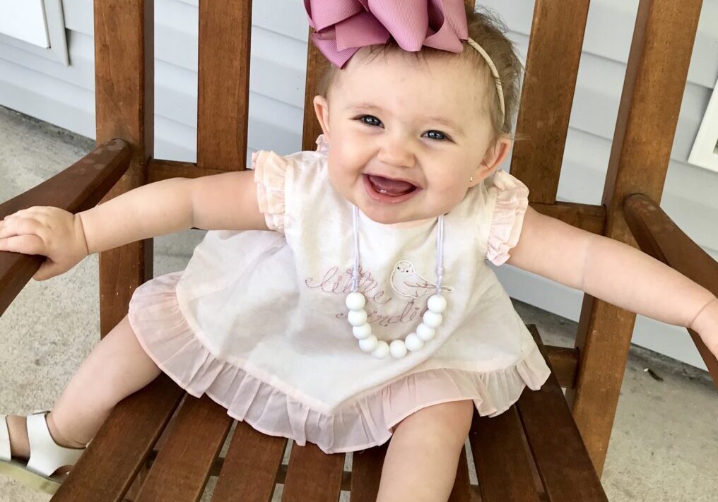 A baby girl sitting on a wooden chair with a pink bow.