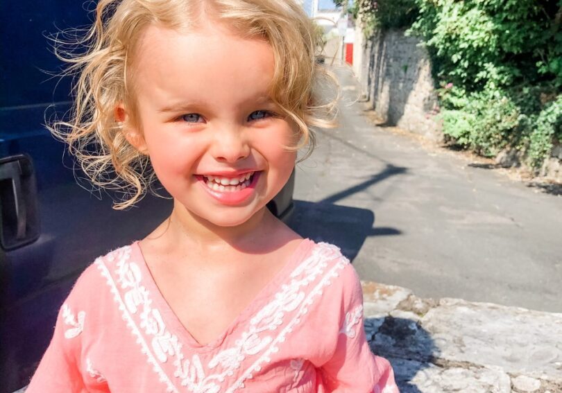 A little girl in a pink dress sitting on a stone wall.