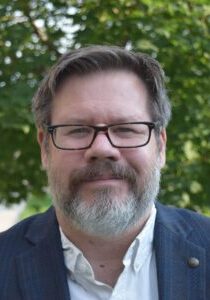 A man with glasses and a beard standing in front of a tree.